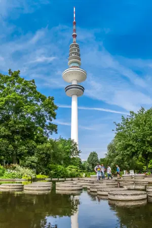 Tall communication tower with distinct circular platforms, surrounded by trees and a clear blue sky. People walk on stepping stones over a small water feature in the foreground.