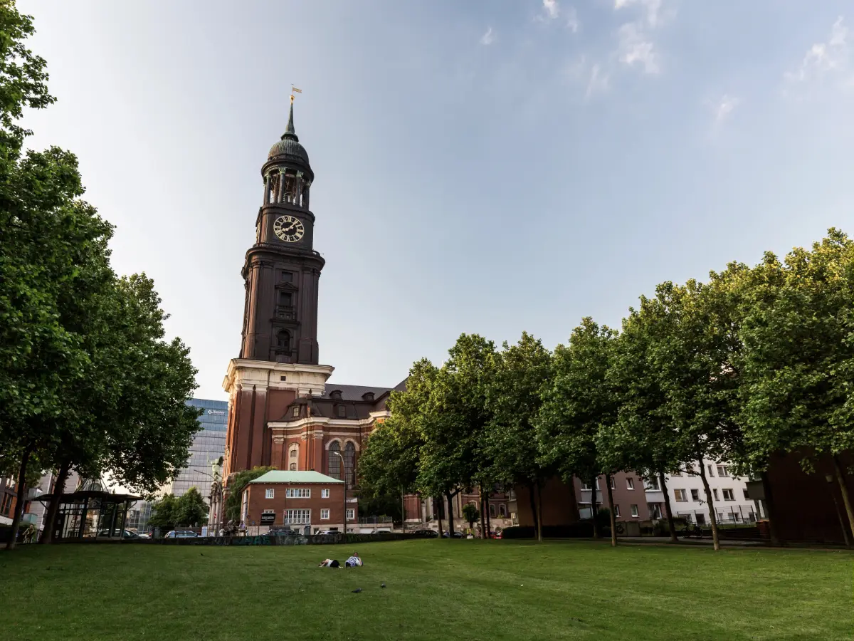 Hamburg's St. Michael's church tower with a clock sits beside a row of trees, overlooking a grassy area under a clear sky.