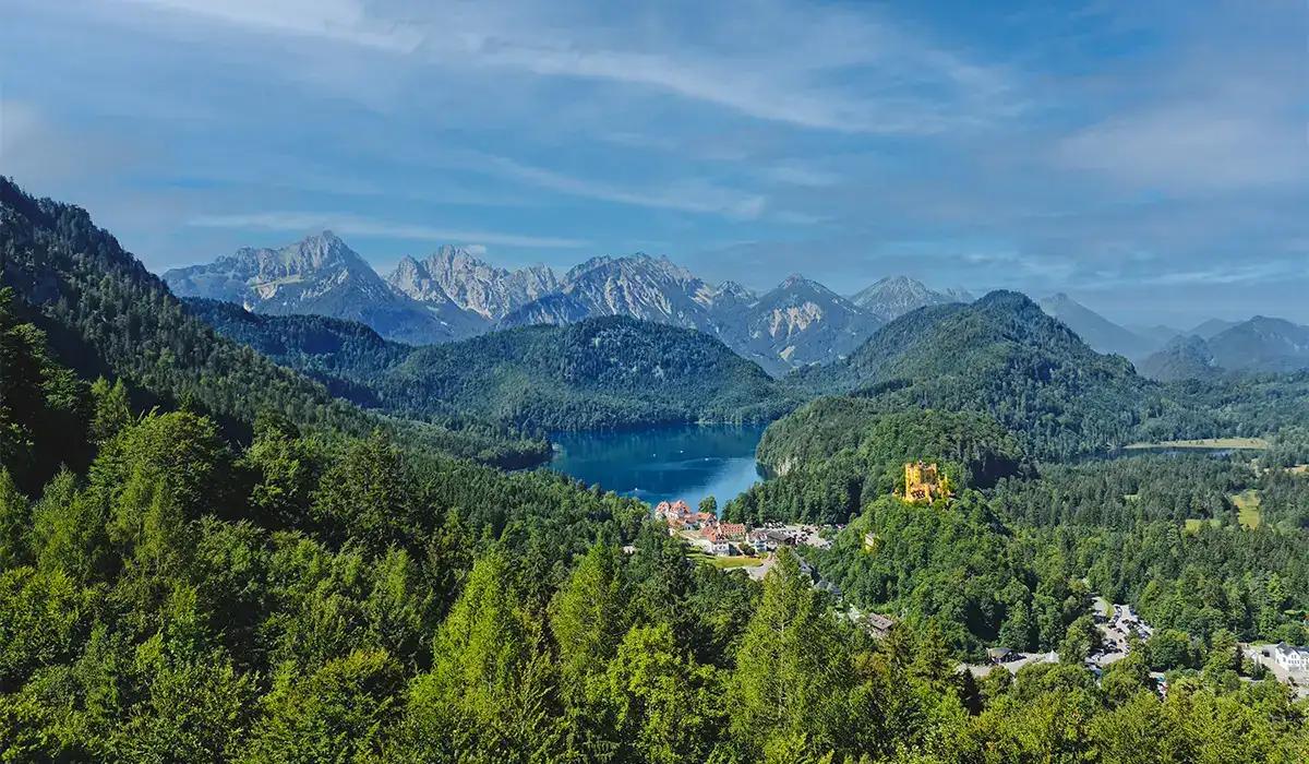 Aerial view of Hohenschwangau Castle and Alpsee near Munich, surrounded by lush forest and the Bavarian Alps.