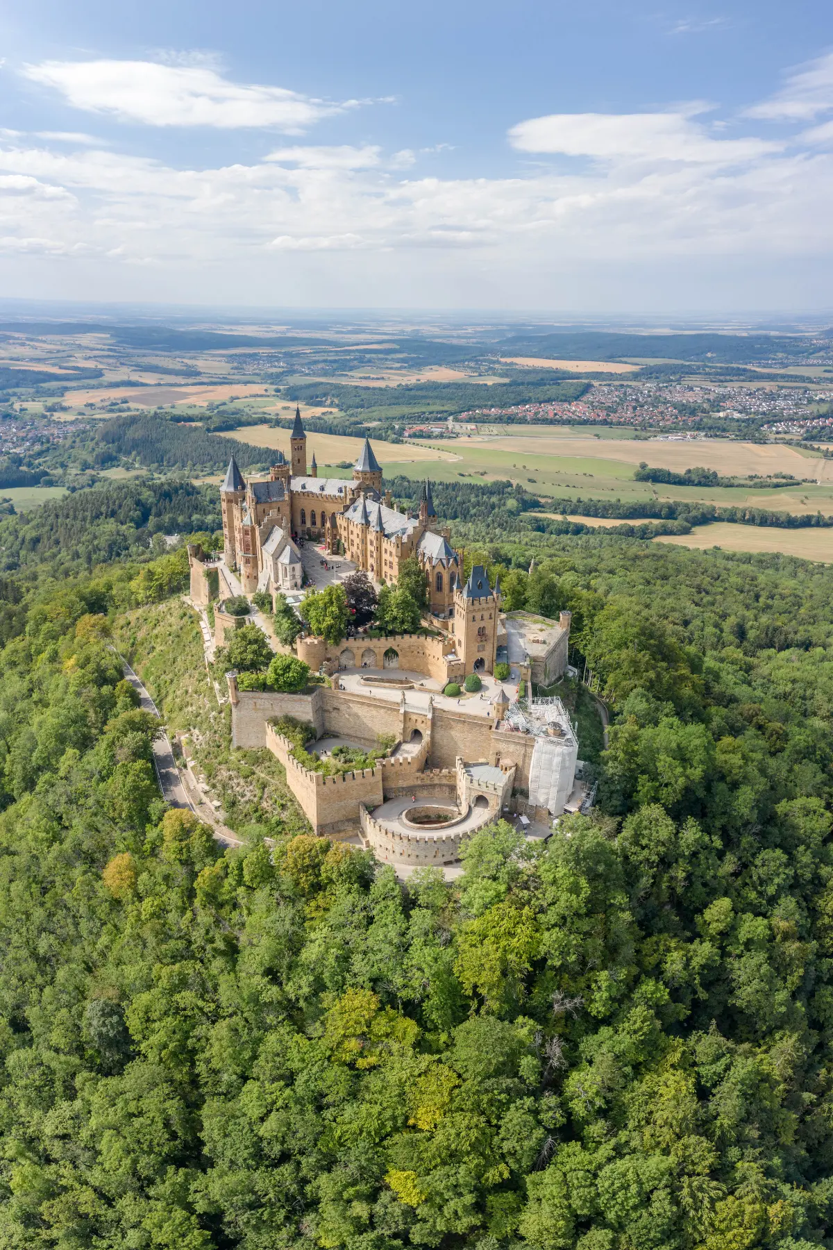 Aerial view of Hohenzollern Castle surrounded by lush greenery, set atop a hill under a partly cloudy sky.