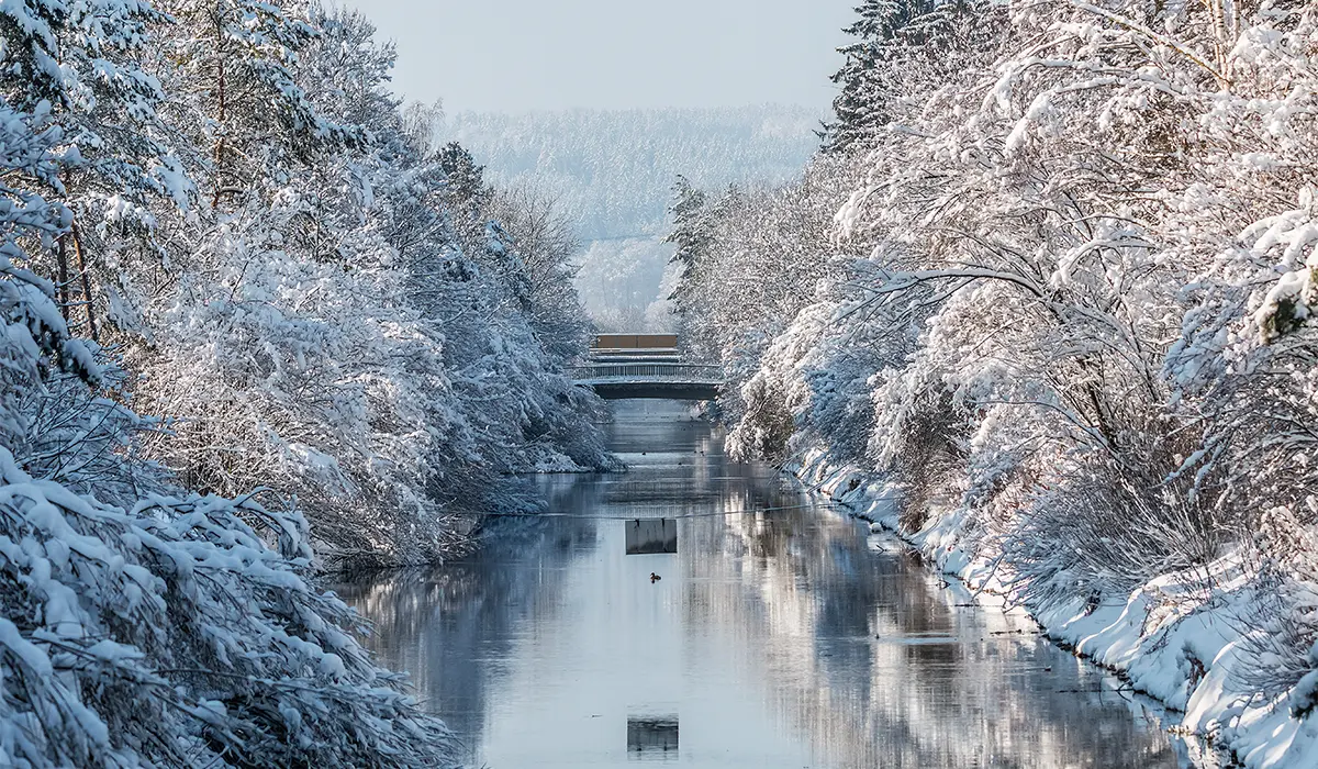 A tranquil snow-covered river scene with trees on both sides, a small bridge in the background, and a partially frozen water surface reflecting the winter landscape.