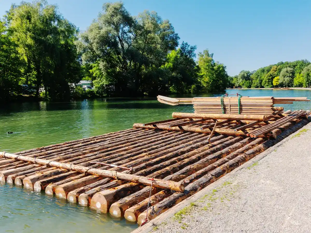 A large wooden raft composed of logs is moored by a riverbank, surrounded by lush greenery under a clear blue sky.