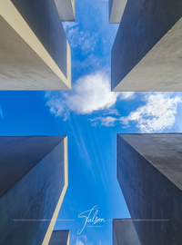 View from below of four tall, rectangular structures at the Memorial for the Murdered Jews in Europe in Berlin, forming a corridor, with blue sky and clouds visible between them.