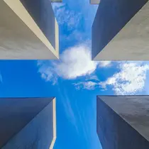 Upward view between the concrete slabs of the Holocaust Memorial in Berlin, framing a blue sky with scattered clouds.