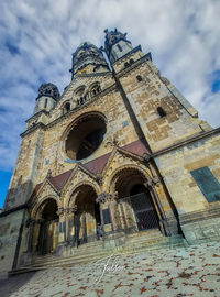 Low-angle view of the Kaiser Wilhelm Memorial Church in Berlin, showing its stone facade, arched entrances, and neo-Romanesque architecture against a partly cloudy sky.