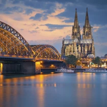 Cologne Cathedral and Hohenzollern Bridge illuminated at dusk, reflected in the calm waters of the Rhine River