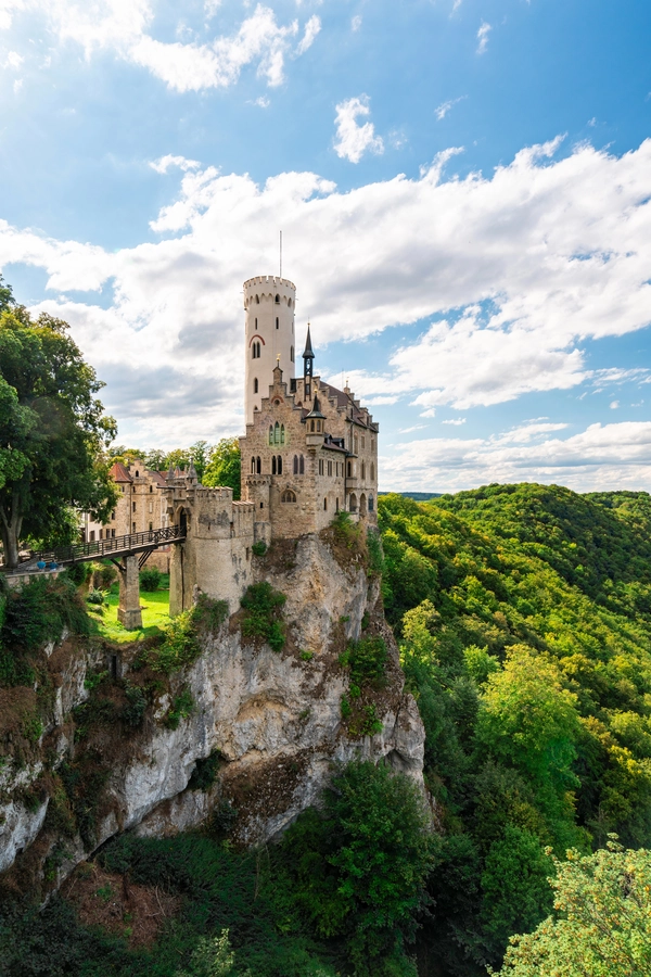 Lichtenstein Castle with a tall tower sits atop a cliff surrounded by lush green trees under a blue sky with scattered clouds.