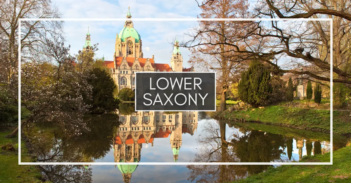 View of a large, historic New Town Hall building in Hannover with green domes and red roofs, reflected in a calm pond, surrounded by trees and grass.