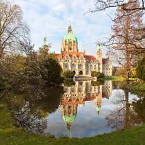 The New Town Hall in Hanover, Germany, reflected in a peaceful pond surrounded by trees in early spring.