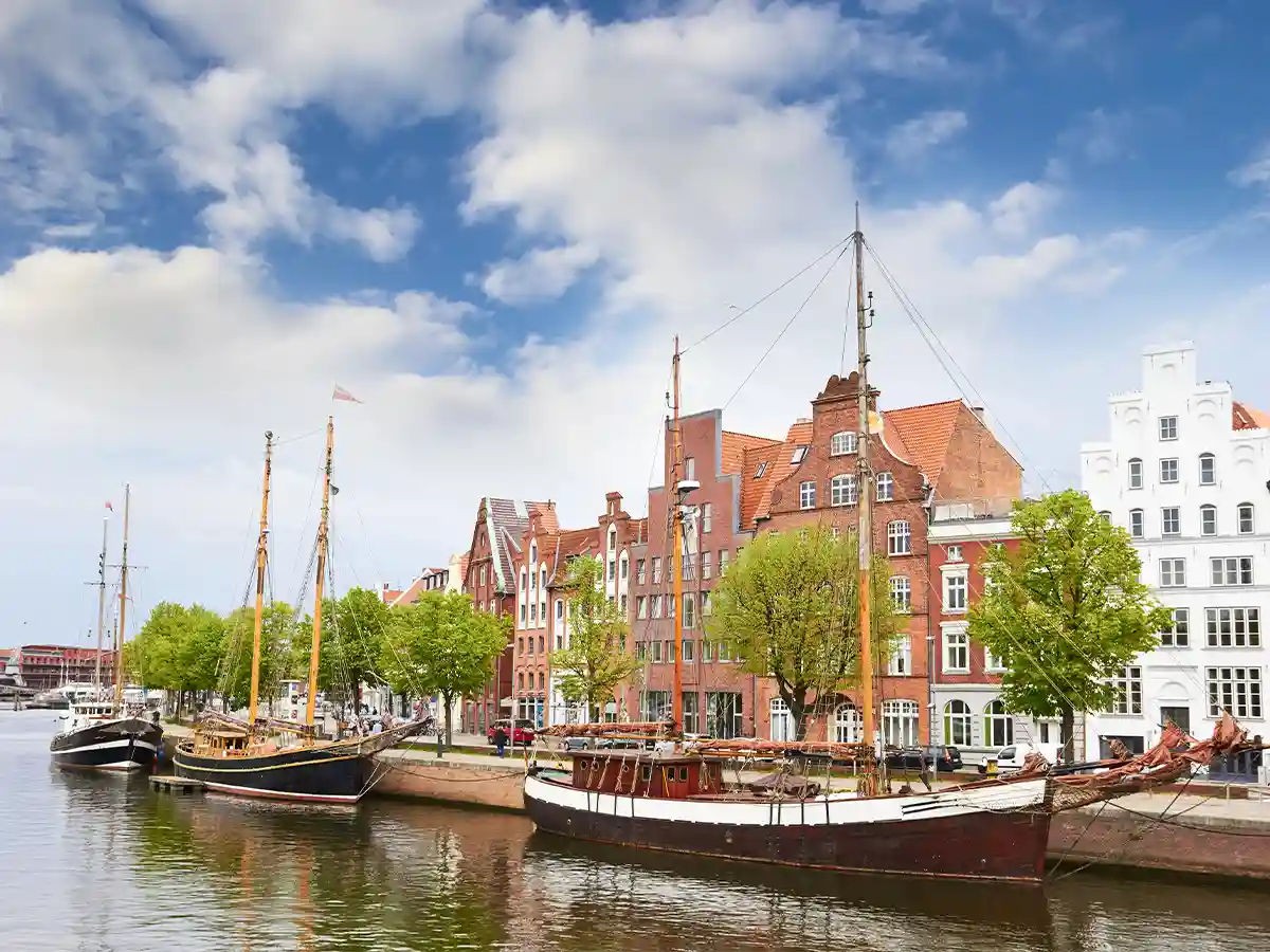 Historic sailing ships docked along a canal with brick and gabled buildings and green trees lining the waterfront under a partly cloudy sky. Historic sailing ships docked along a canal with brick and gabled buildings and green trees lining the waterfront under a partly cloudy sky.