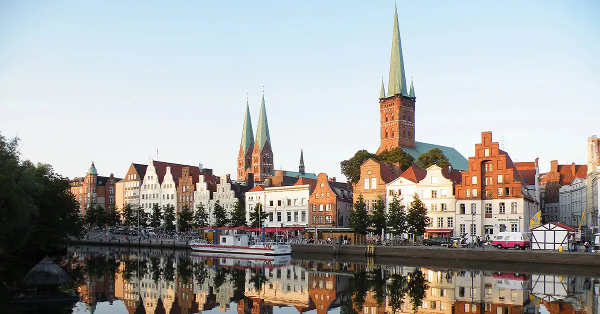 Historic skyline of Lübeck, Germany, with stepped-gable houses, spired churches, and a tour boat reflected in the Trave River during golden hour.