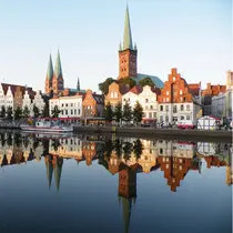 Historic skyline of Lübeck, Germany, with stepped-gable houses, spired churches, and a tour boat reflected in the Trave River during golden hour.
