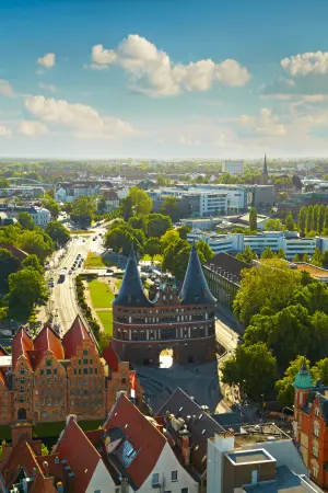 Aerial view of Lübeck, Germany, featuring the Holstentor gate flanked by lush greenery and historic red-roofed buildings under a blue sky.