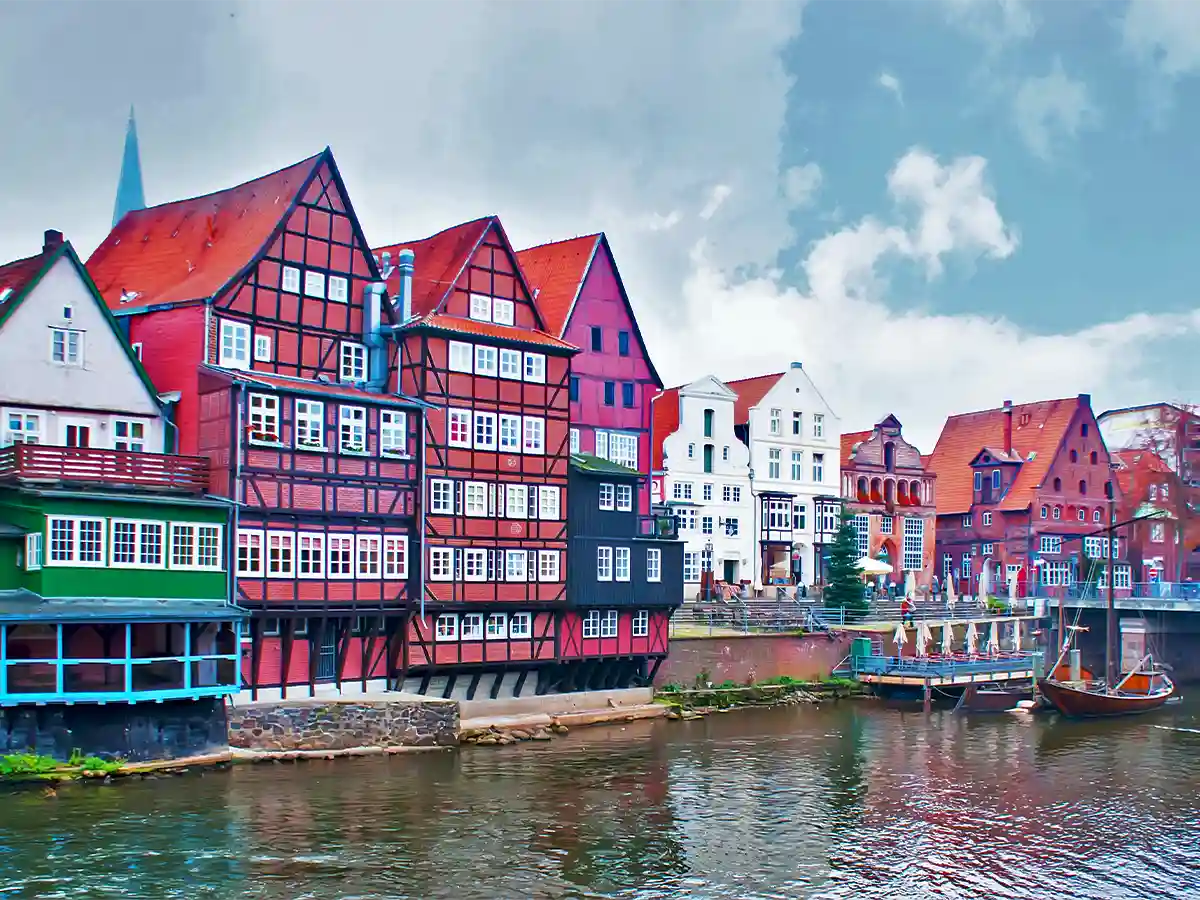 Colorful historic buildings with timber framing line a riverbank under a cloudy sky, with boats docked along the water in Lüneburg. Colorful historic buildings with timber framing line a riverbank under a cloudy sky, with boats docked along the water in Lüneburg.
