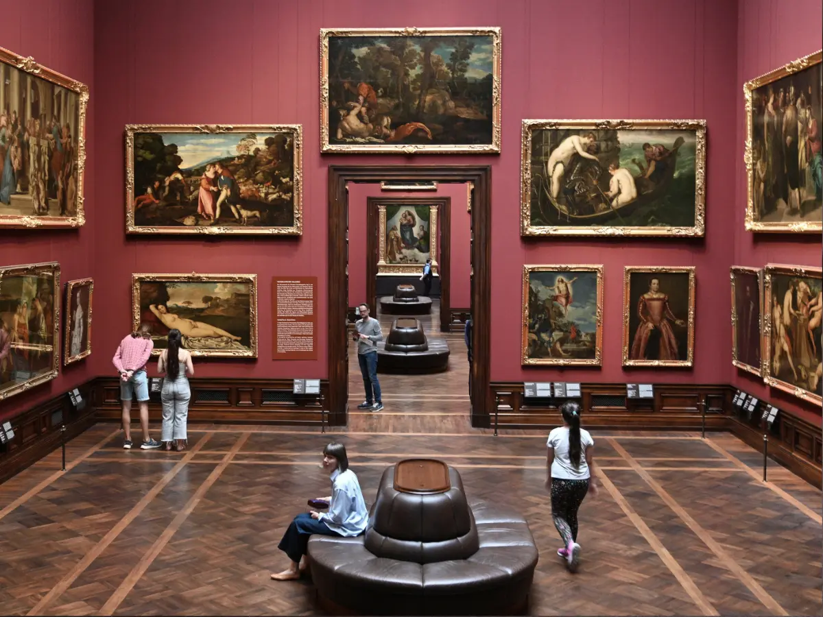 Masters Art Gallery in Dresden Dresden art museum gallery with visitors viewing paintings on red walls. Central hallway leads to more artwork. Some people sit on circular benches on a wooden floor.