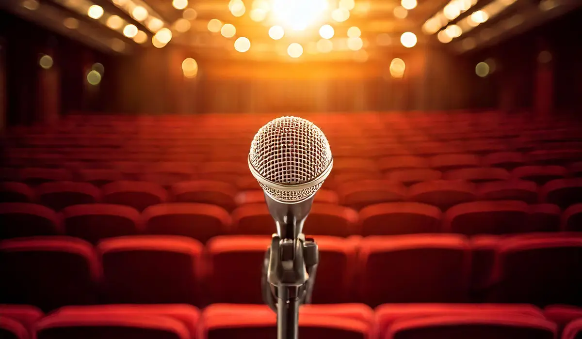 A microphone on a stand faces an empty theater with red seats and bright stage lights in the background.