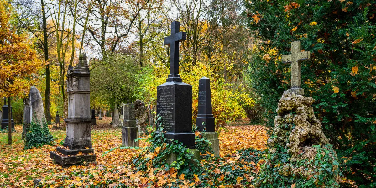Historic gravestones in the Old North Cemetery of Munich, Germany surrounded by fallen leaves and trees.