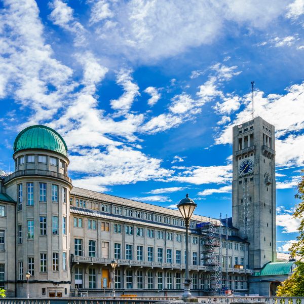 The Munich Deutsches Museum with grey building dotted with windows, clock tower, and curved tower with green domed roof, against a bright blue sky dotted with clouds