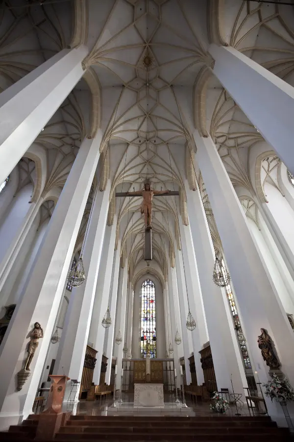 Interior view of the Munich Frauenkirche with tall white columns, a large cross hanging from the ceiling, stained glass windows, and a stone altar.