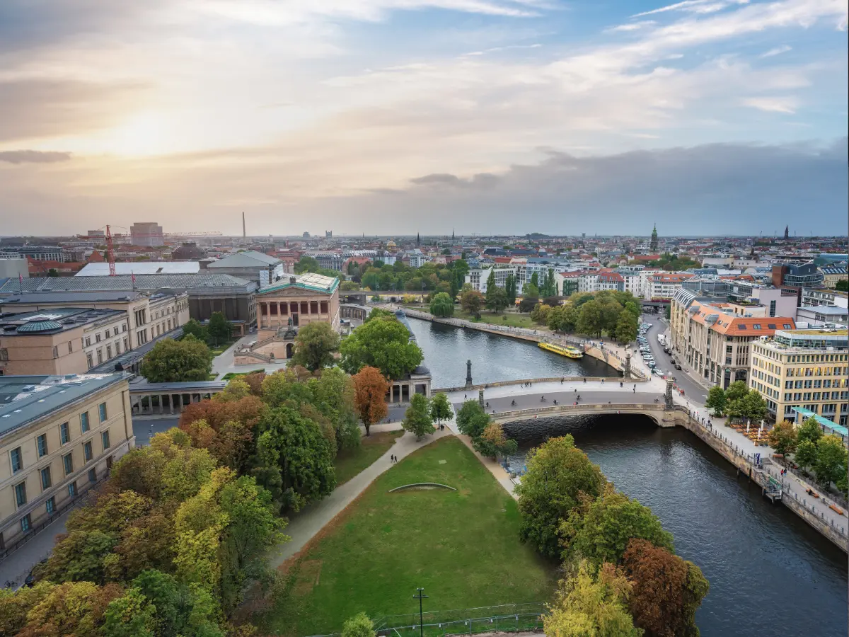 Museum Island in Berlin Aerial view of a cityscape showing a river winding through buildings and green spaces on Museum Island in Berlin, with bridges crossing over. The sky is partly cloudy with a sunset in the background.