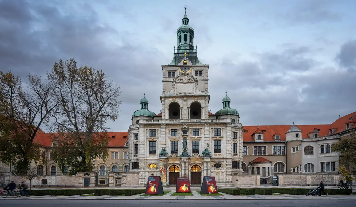 Front view of the Deutsches Museum building with copper domes and ornate stone architecture under a cloudy sky.