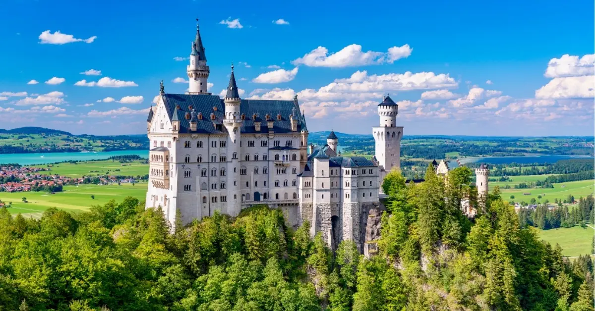 Neuschwanstein Castle perched above green forested hills in Bavaria, with clear blue skies and distant lakes creating a postcard-perfect fairytale scene.