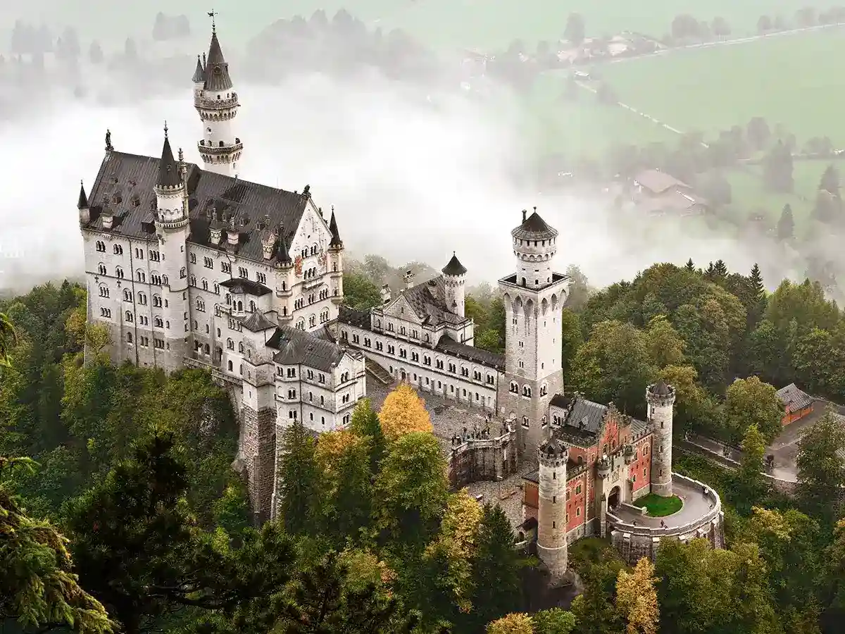 Neuschwanstein Castle sits atop a wooded hill, surrounded by mist and greenery, with towers and spires creating a fairytale-like appearance.