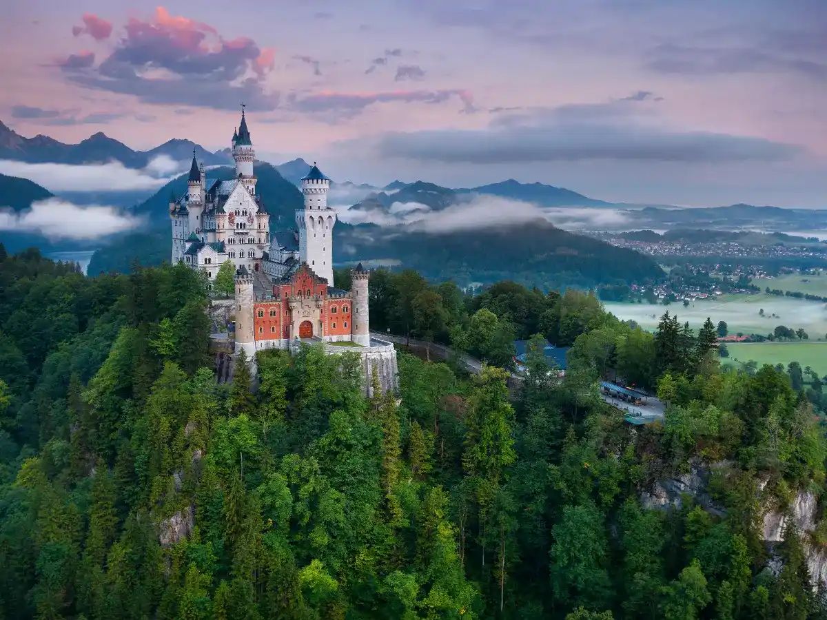 Aerial view of Neuschwanstein Castle atop a forested hill, surrounded by mist and mountains in the background, under a cloudy sky during twilight.
