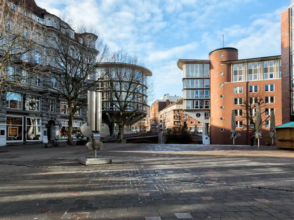A paved city square with a modern metal sculpture in the center, surrounded by contemporary and older brick buildings under a blue sky. A paved city square with a modern metal sculpture in the center, surrounded by contemporary and older brick buildings under a blue sky.
