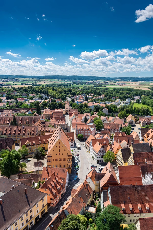 Aerial view of Nördlingen town with red-roofed buildings and a distinctive triangular structure, surrounded by greenery and fields under a blue sky with clouds.