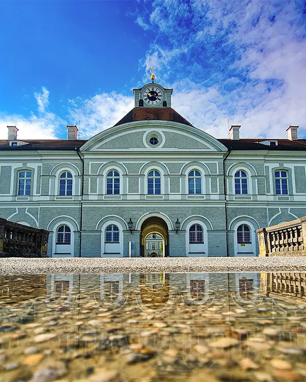 Symmetrical view of Nymphenburg Palace entrance with arched windows and a reflection in a shallow puddle.