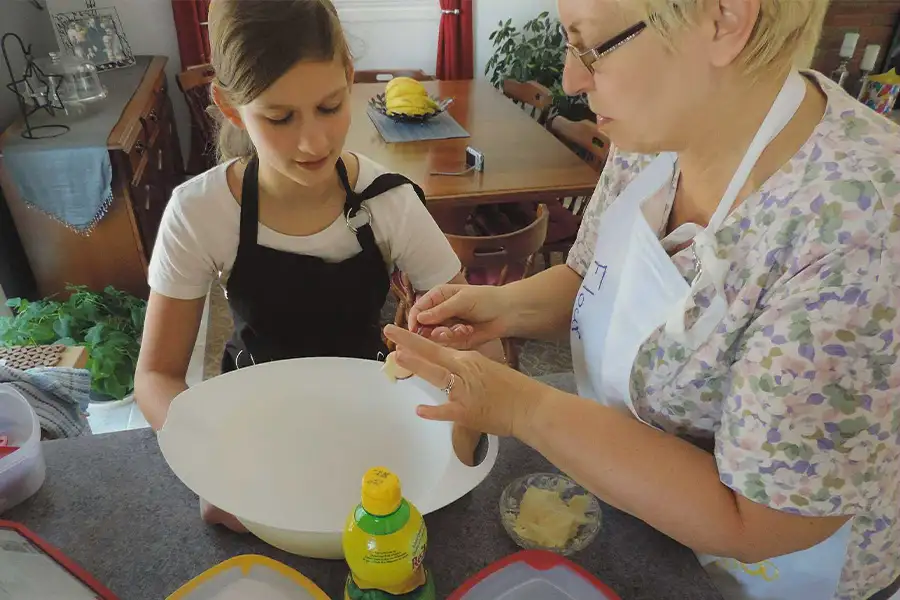 Grandmother teaching her granddaughter to bake in a kitchen, both wearing aprons and working together over a large mixing bowl.