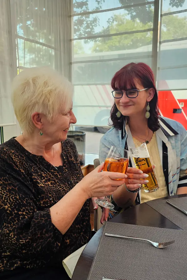 Two women seated at a table clink their drinks in a toast. One woman holds a glass of wine, and the other holds a beer. They are smiling and appear to be enjoying their time together.