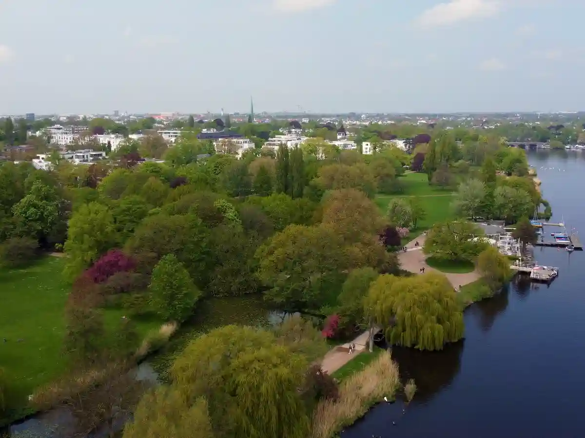Aerial view of a lush green park with walking paths, trees, and a body of water, with a cityscape visible in the background. Aerial view of a lush green park with walking paths, trees, and a body of water, with a cityscape visible in the background.