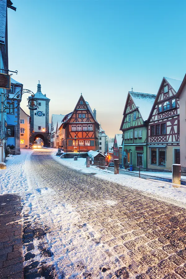 Snow-covered cobblestone street lined with colorful half-timbered buildings under a clear blue sky in Rothenburg, Germany.