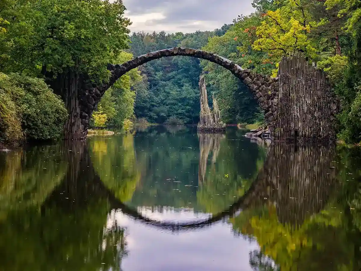 Arched stone bridge over a calm, reflective lake, surrounded by dense green foliage in a forest setting.