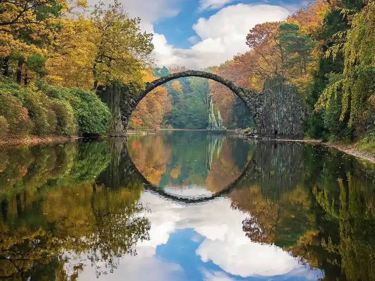 Stone arch bridge over a calm lake with a perfect circular reflection. Surrounded by autumn trees under a partly cloudy sky.