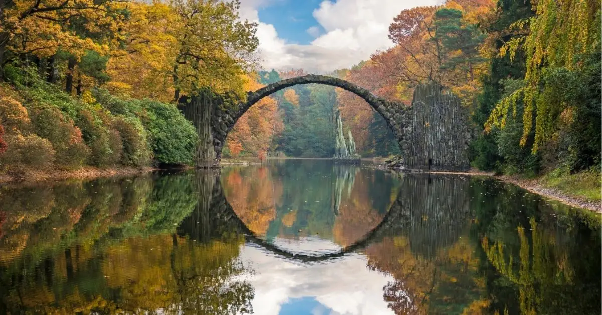 Rakotzbrücke (Devil’s Bridge) in Saxony, Germany, surrounded by vibrant autumn foliage and reflected perfectly in the still water below, forming a complete stone circle.