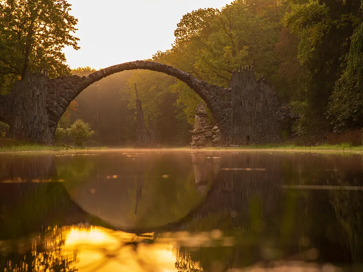 Stone arch bridge with reflection on a calm river, surrounded by lush green trees under a warm golden sky.