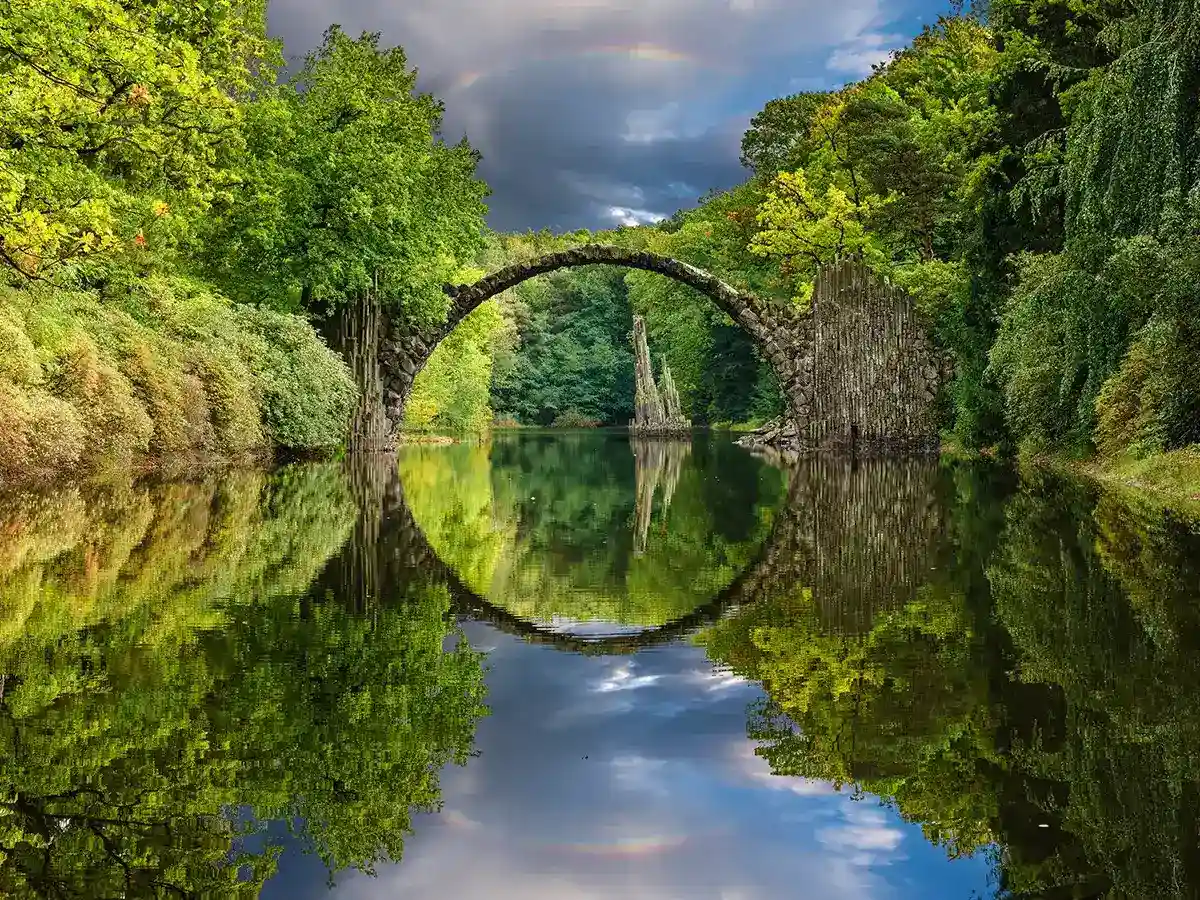 Stone bridge arches over a calm river, surrounded by lush green trees, with its reflection creating a circle on the water. Cloudy sky above.