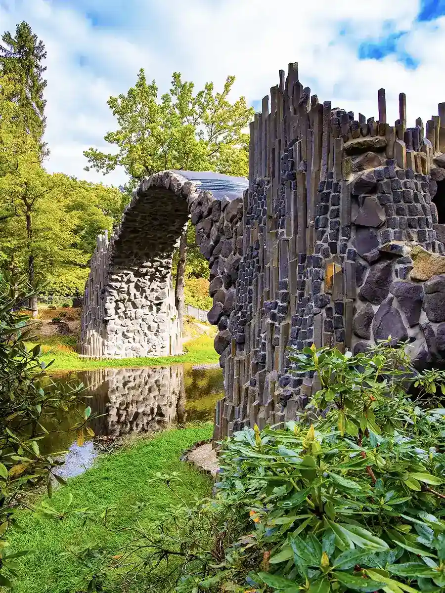 Stone bridge with an arch made of columnar basalt reflected in a pond, surrounded by greenery and trees under a partly cloudy sky.
