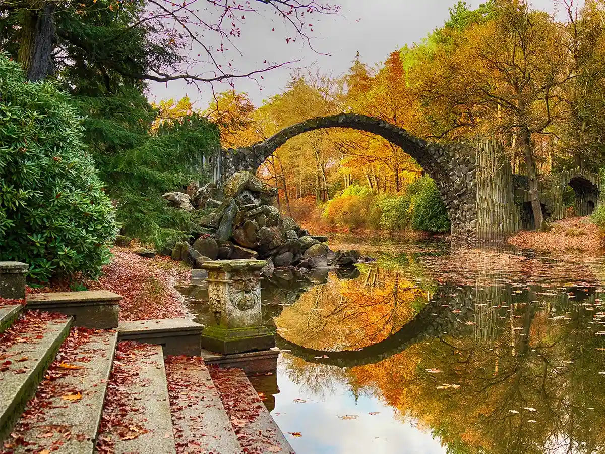 Stone arch bridge with reflection in a calm river, surrounded by autumn trees and fallen leaves on stone steps in the foreground.