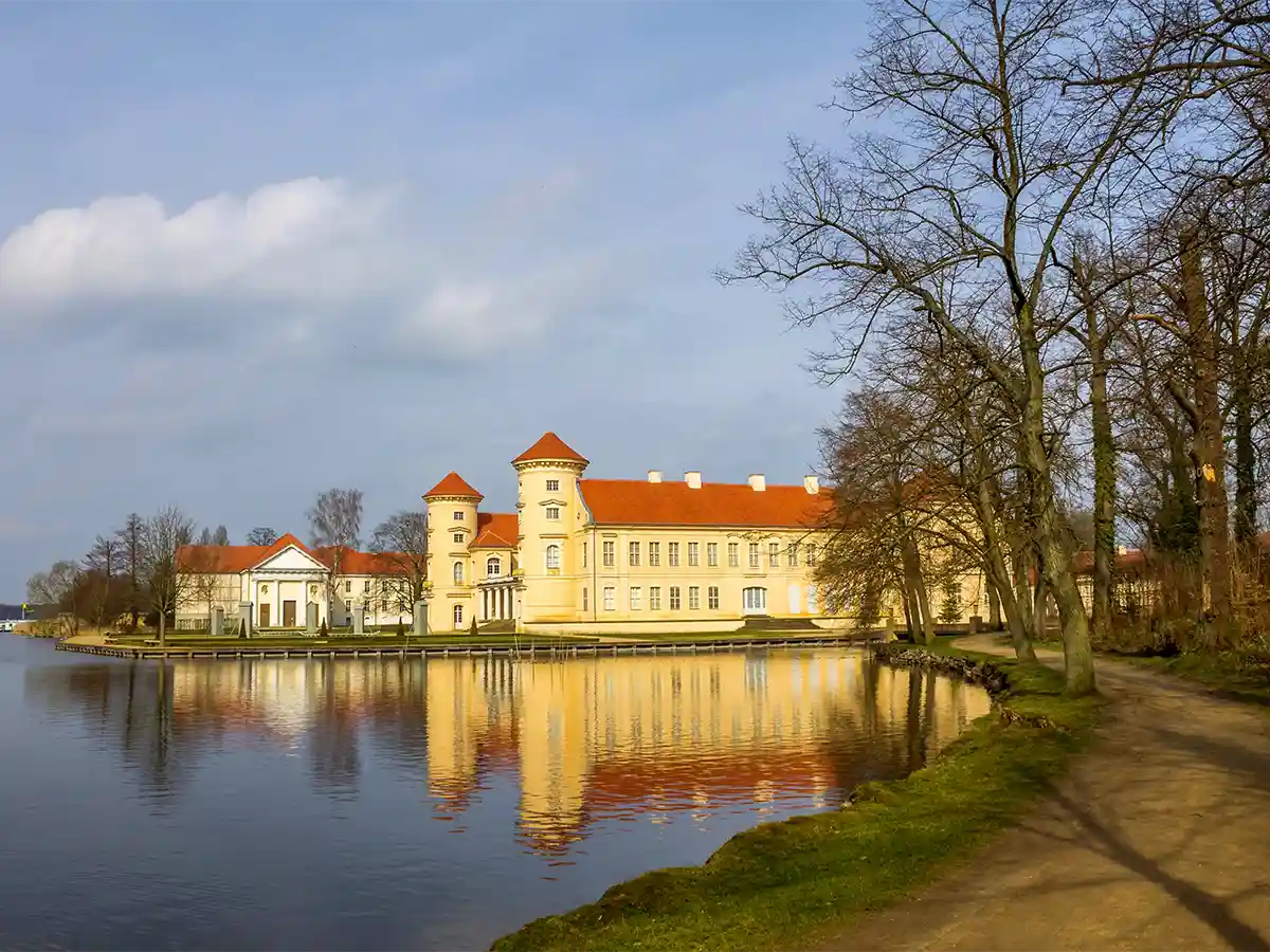 Lake-front Rheinsberg Castle glowing in afternoon sun, reflection rippling—must-see Brandenburg castles and lakes.