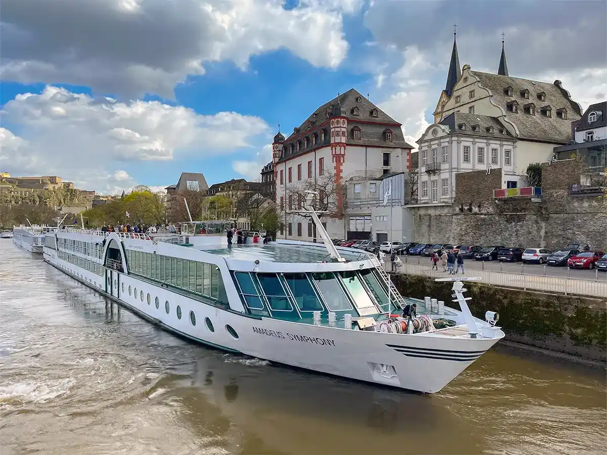 River cruise ship "Amadeus Symphony" navigates through a channel with historic buildings and cars lining the waterfront. Cloudy sky above.
