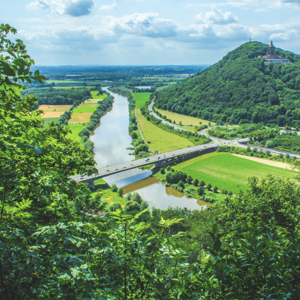 View from a hill overlooking a winding river, intersected by a bridge. Lush green fields and dense forests are seen with a building on a hilltop in the distance under a partly cloudy sky.