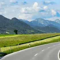Scenic winding road cutting through green meadows and alpine mountains under a partly cloudy sky
