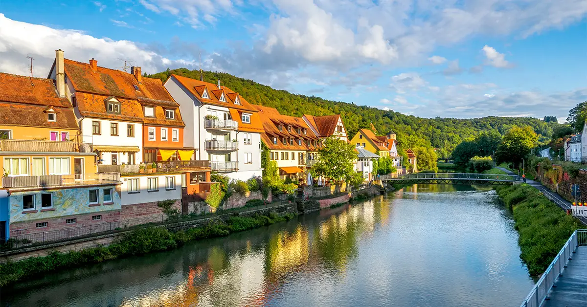 Scenic riverside view of colorful timbered houses and modern buildings in Wertheim, Germany, with a lush green forest in the background and a small bridge crossing the river.