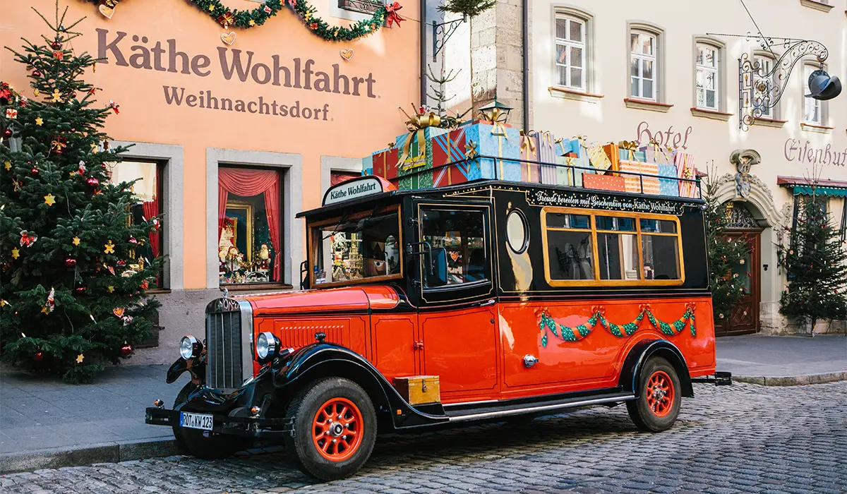 Vintage red and black bus decorated with garlands and gifts on the roof, parked next to a building with Christmas trees and festive signage.