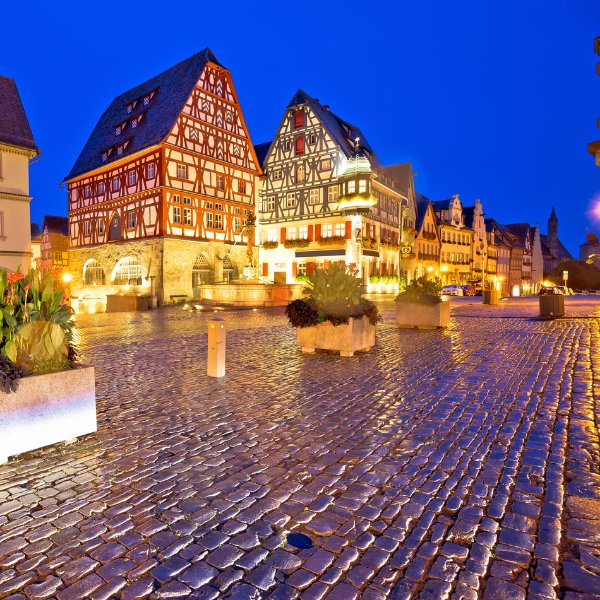 Rothenburg at Christmas A cobblestone square with illuminated traditional half-timbered buildings at dusk, featuring flower pots and a clear blue sky.