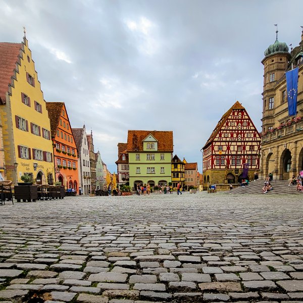 A cobblestone town square with colorful half-timbered buildings under a cloudy sky. People can be seen walking and sitting.